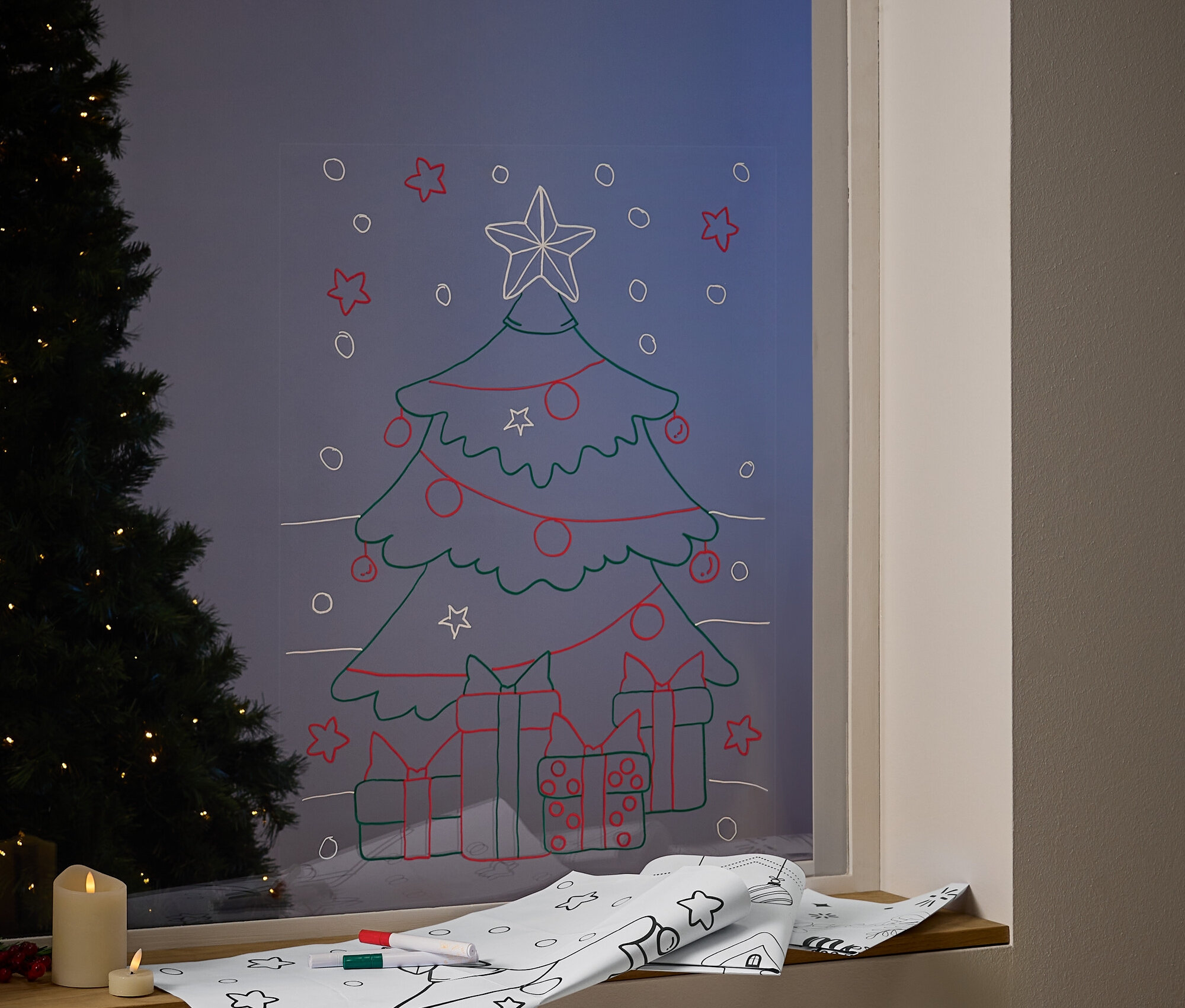 Na oknie narysowana jest choinka z ozdobami i prezentami. Na parapecie leżą papier z motywami świątecznymi i pisaki.
