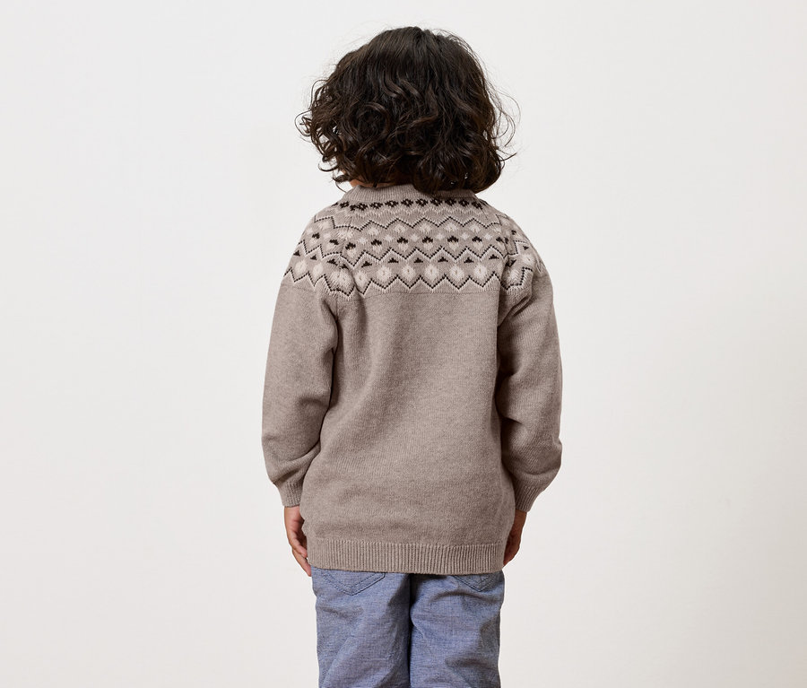 Chłopiec z ciemnymi kręconymi włosami stoi tyłem do kamery, ubrany w brązowy sweter we wzory i niebieskie spodnie.