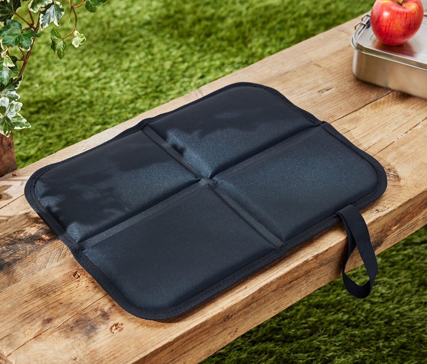Składana poduszka do siedzenia z izolacją termiczną leży na drewnianej ławce obok jabłka i pudełka na lunch.