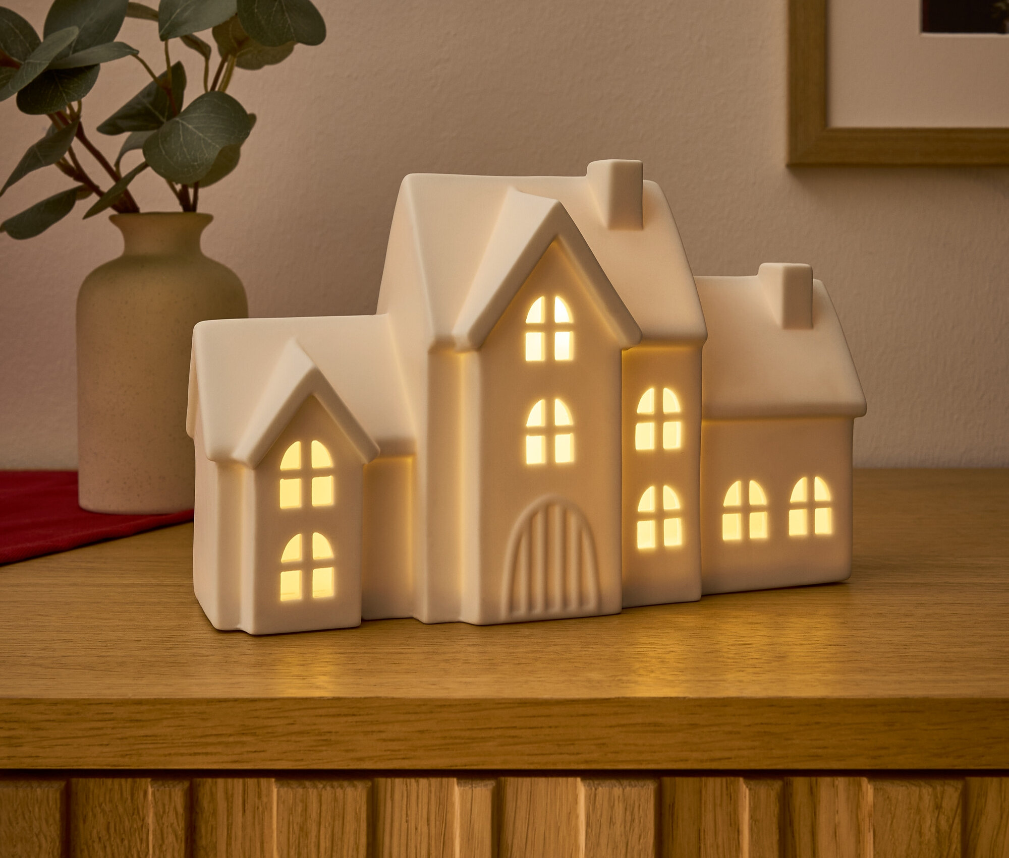 Na drewnianej komodzie stoi ceramiczny, świecący dom i wazon z eukaliptusem.