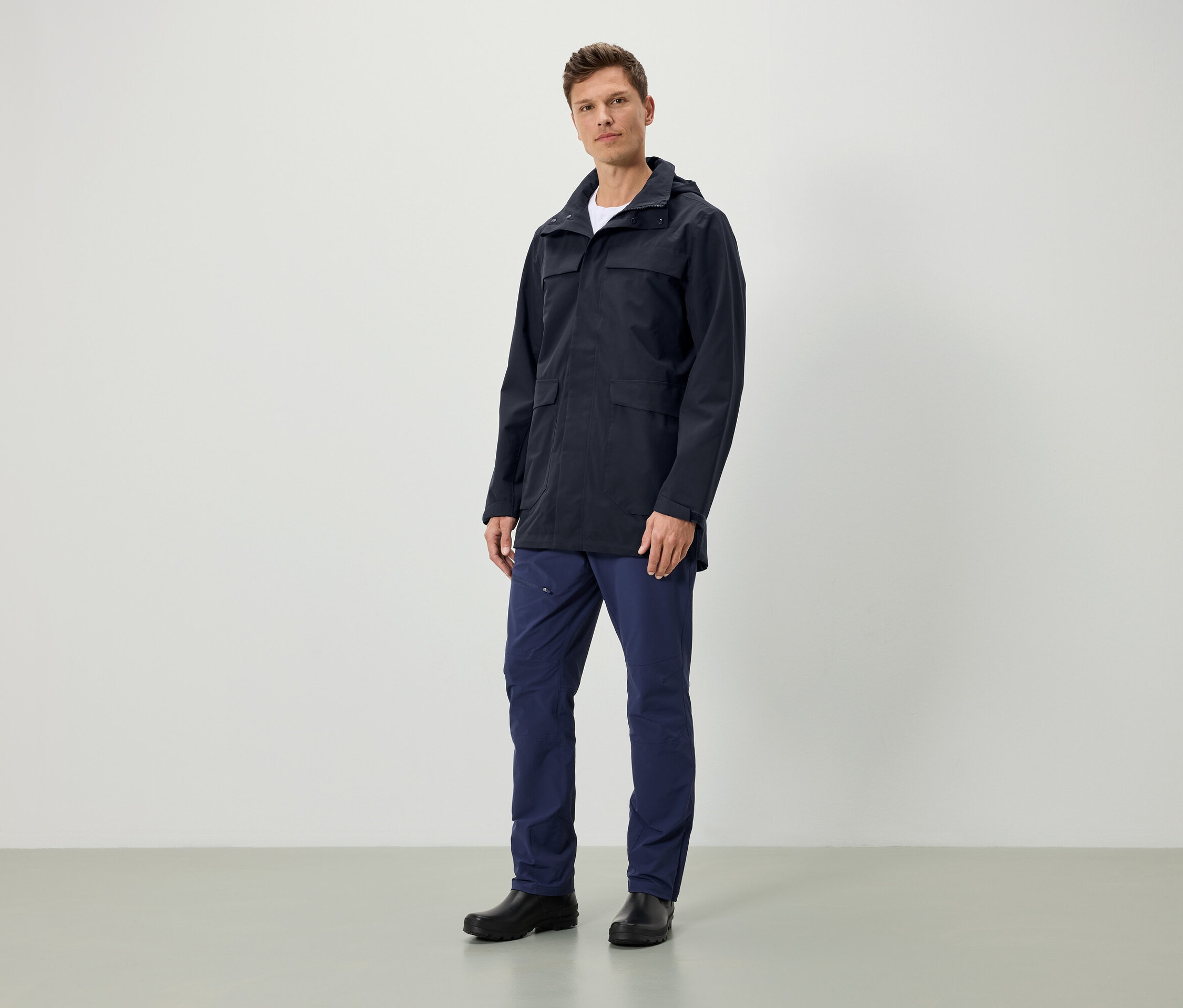 Mężczyzna stoi i ma na sobie granatową kurtkę z kapturem, granatowe spodnie i czarne buty.