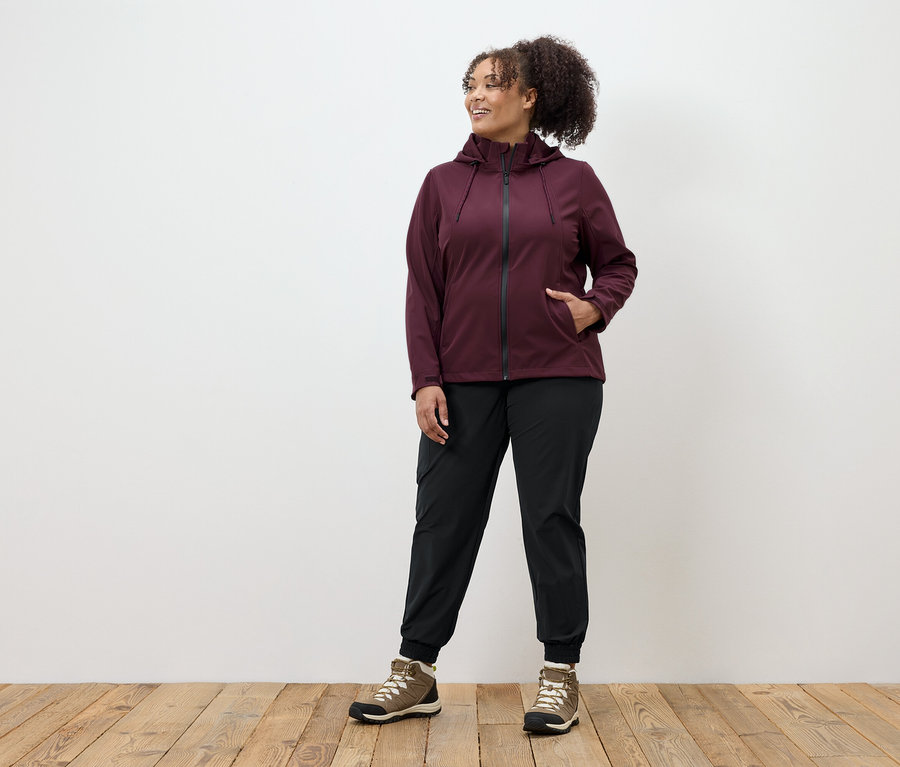 Kobieta w bordowej kurtce na zamek błyskawiczny, czarnych spodniach i brązowych butach stoi na drewnianej podłodze.