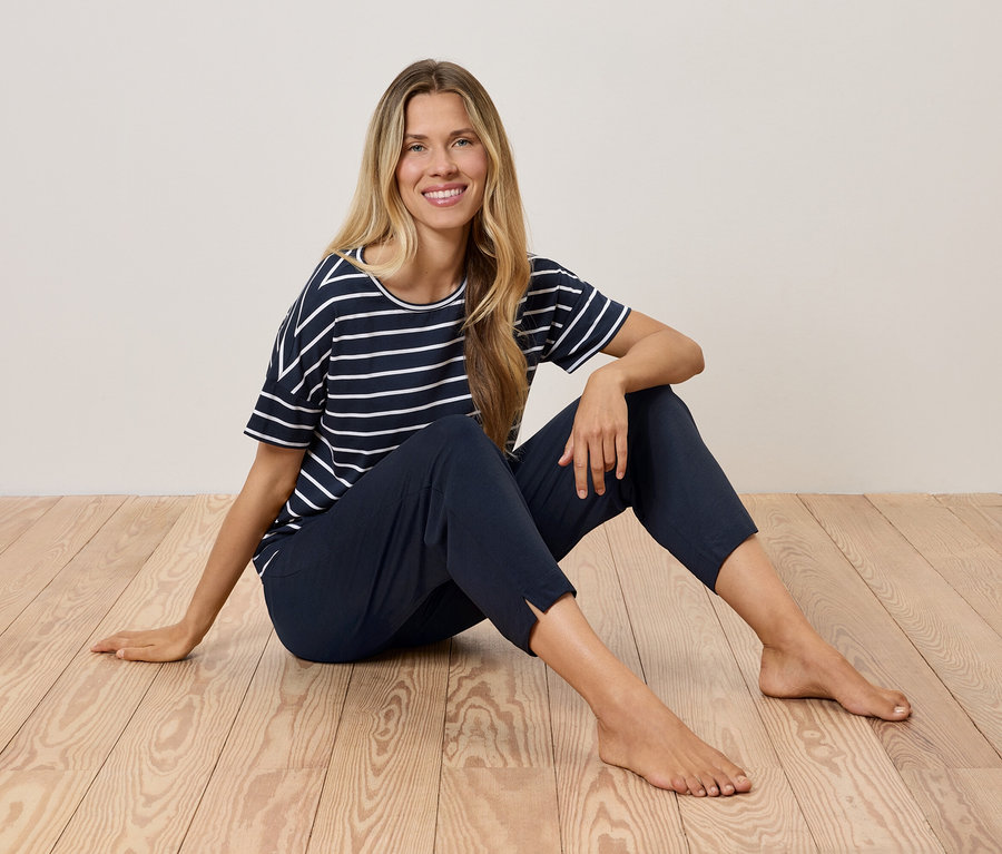 Blondynka siedzi na drewnianej podłodze w granatowo-białej koszulce w paski i granatowych spodniach.