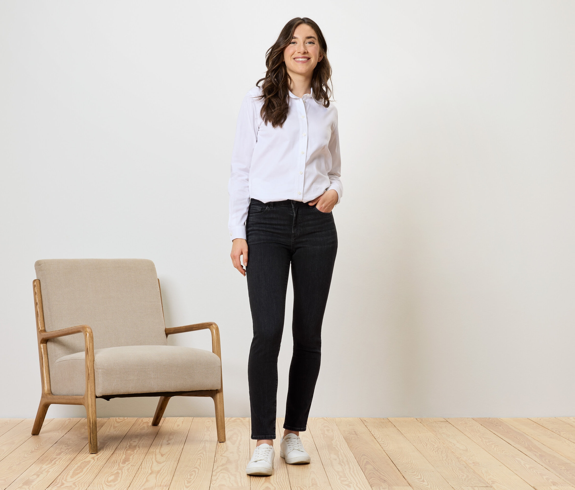 Kobieta stoi w czarnych dżinsach, białej koszuli i białych butach obok beżowego krzesła.