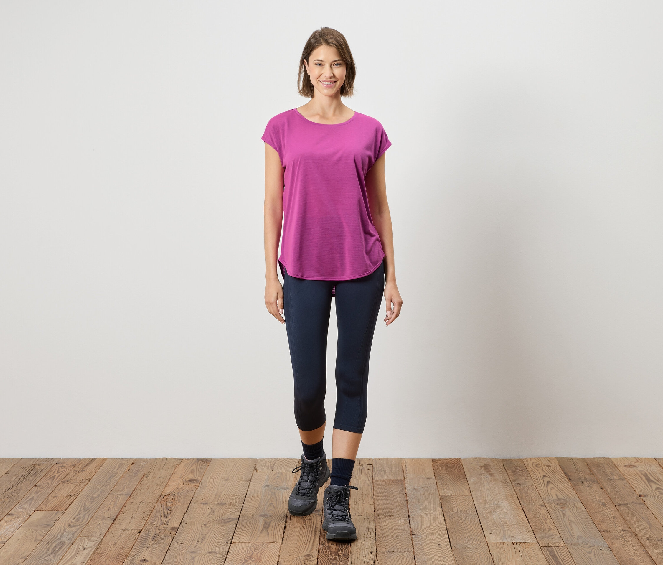 Kobieta stoi na drewnianej podłodze na tle białej ściany. Ma na sobie różowy T-shirt, niebieskie legginsy i szare buty.