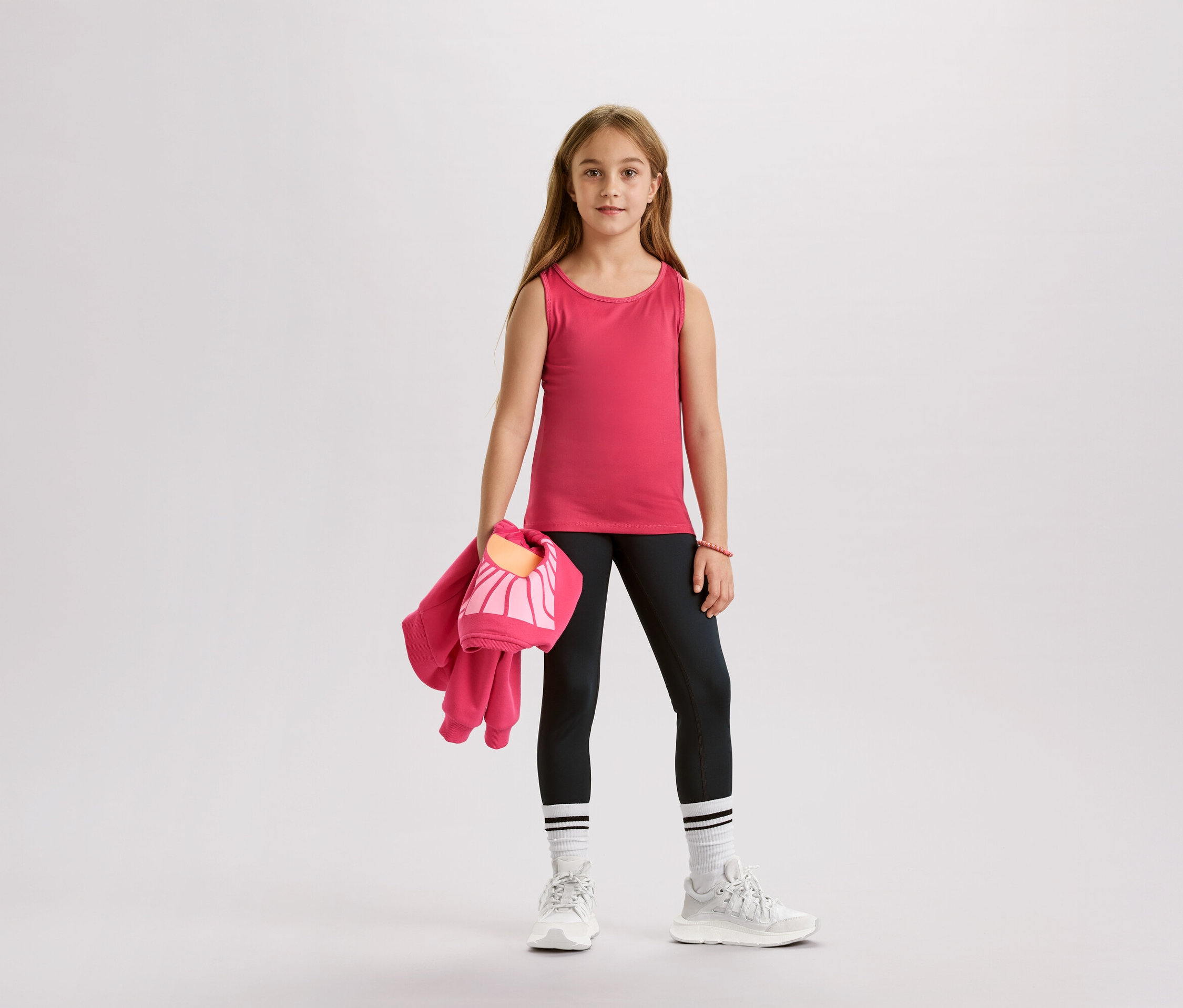 Dziewczyna w różowym topie, czarnych legginsach, białych skarpetach i butach sportowych, trzyma różową kurtkę i torbę.