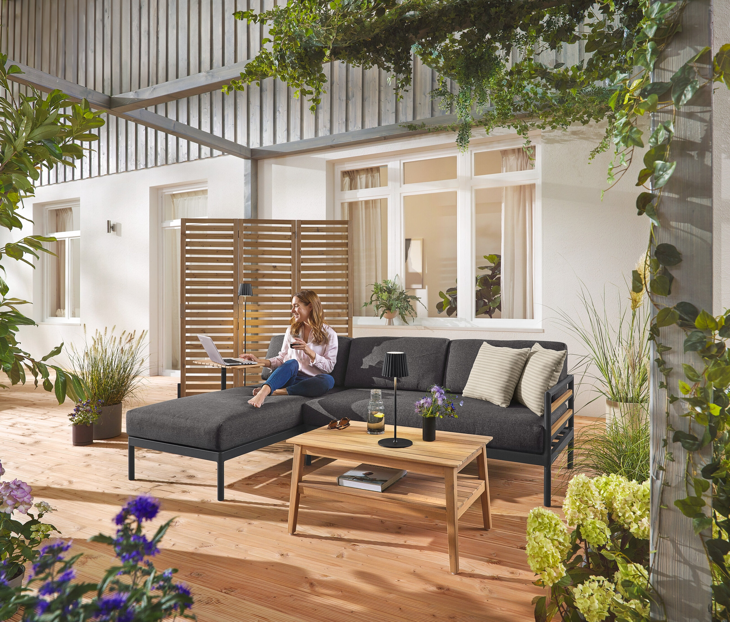 Kobieta siedzi na narożniku ogrodowym Premium »Elin«, w tle znajduje się elastyczny parawan. Widoczna jest również czarna, akumulatorowa lampa podłogowa i stołowa LED oraz stolik kawowy.