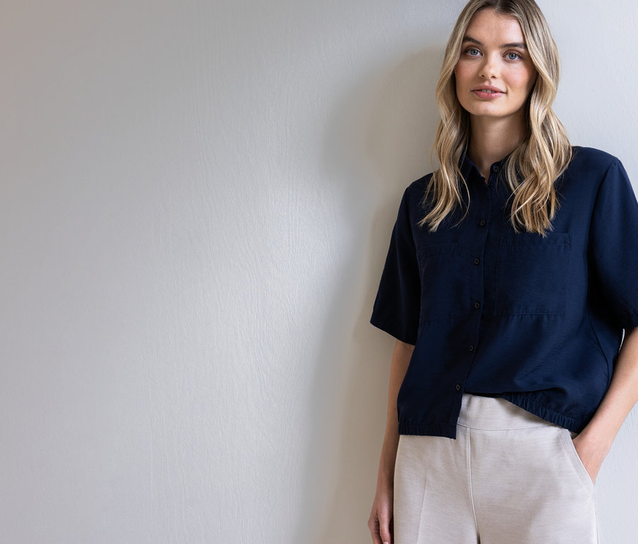 Kobieta o blond włosach w ciemnoniebieskiej koszuli i beżowych spodniach pozuje na tle jasnoszarej ściany.