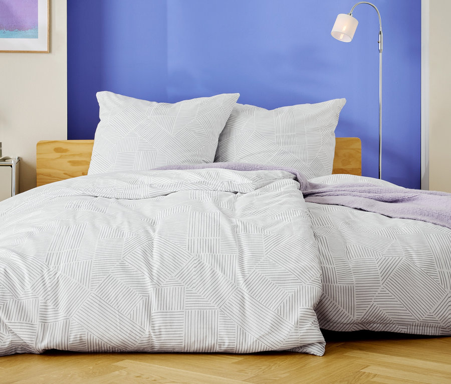 Łóżko z białą pościelą w geometryczne wzory i dwiema poduszkami stoi na tle niebieskiej ściany.