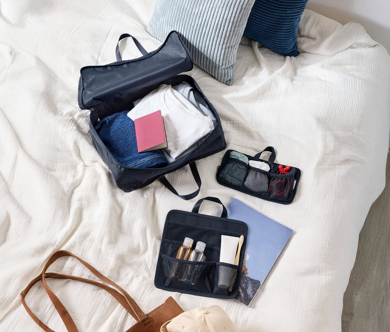 Widok z góry na walizkę z ubraniami, dwie poduszki, kosmetyczkę, torbę na ramię i czapkę na łóżku.