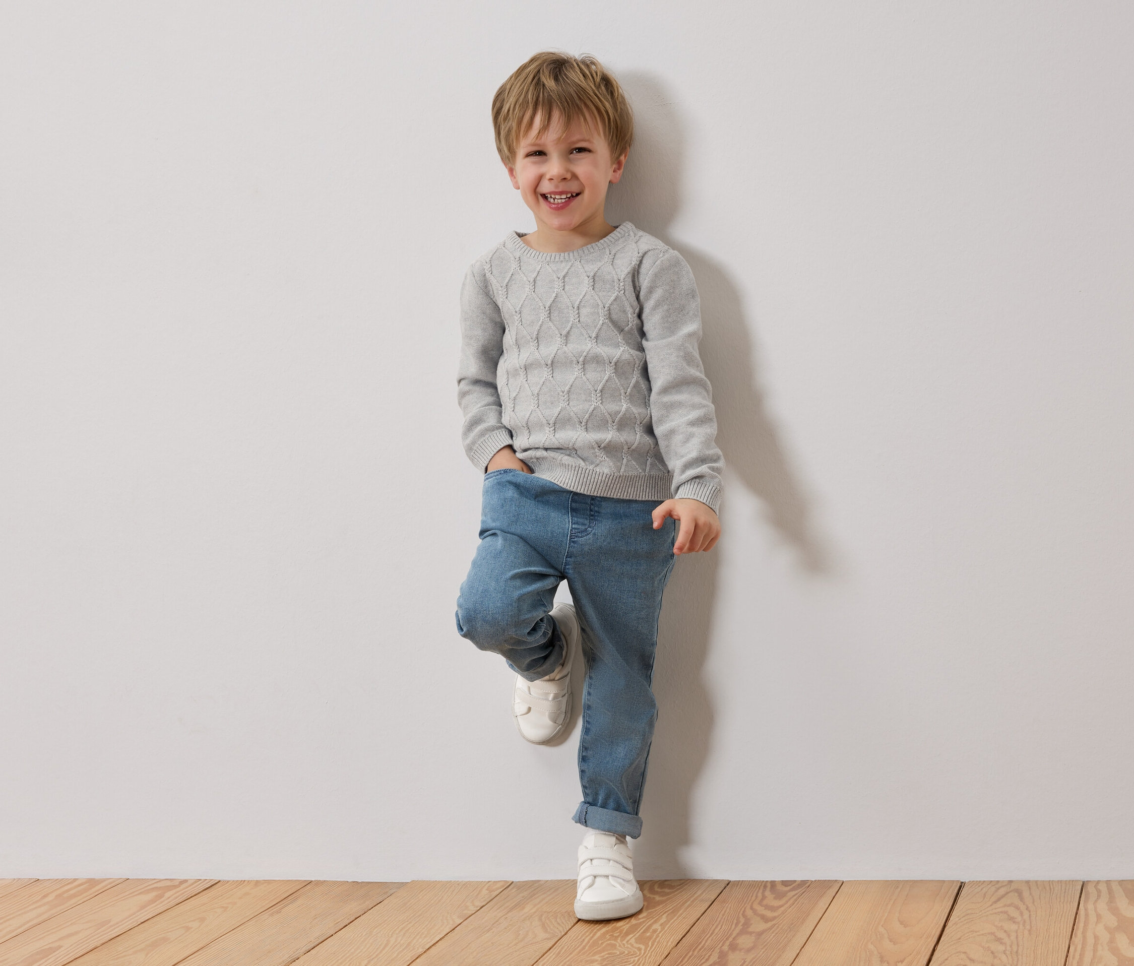 Uśmiechnięty blond chłopiec opiera się o białą ścianę. Ma na sobie szary sweter, niebieskie dżinsy i białe buty.