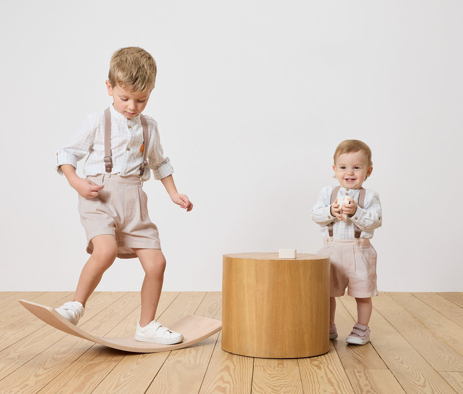 Dwoje dzieci, chłopiec balansuje na drewnianej desce, niemowlę stoi obok drewnianego stolika i trzyma klocek.