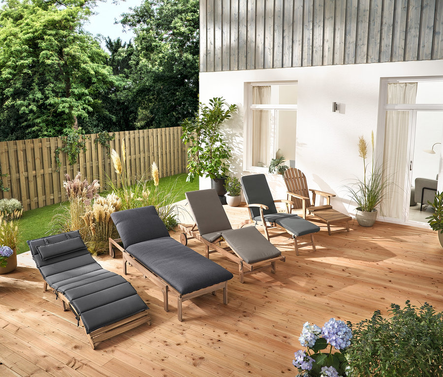 Na drewnianym tarasie stoi kilka leżaków ogrodowych i fotel ogrodowy typu Adirondack z wysuwanym podnóżkiem wykonany z drewna.