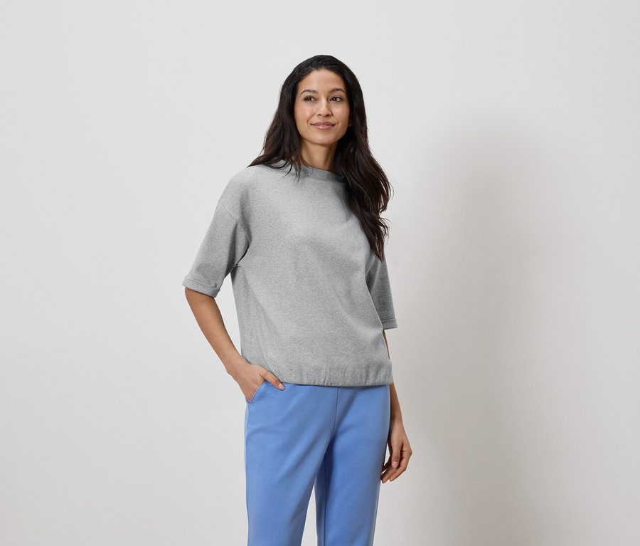 Kobieta o ciemnych włosach ma na sobie szary top i niebieskie spodnie, ręka w kieszeni.