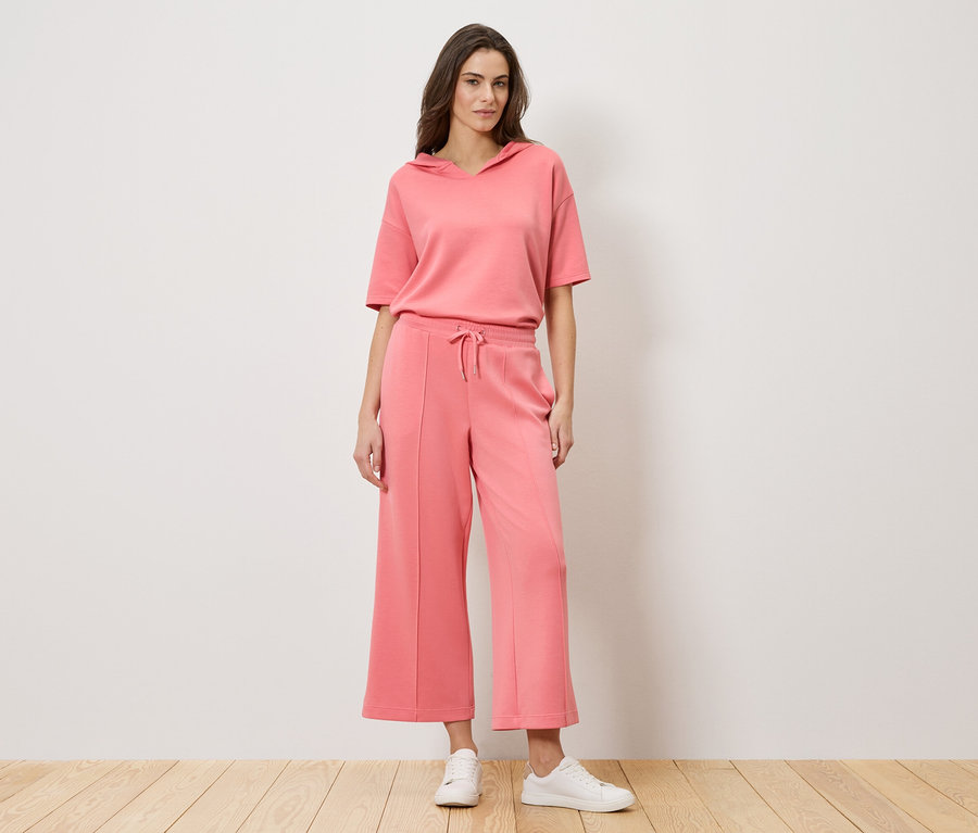 Kobieta stoi na tle ściany, ubrana w koralowy dwuczęściowy strój składający się z bluzy z krótkim rękawem i spodni typu culottes, oraz białych tenisówek.