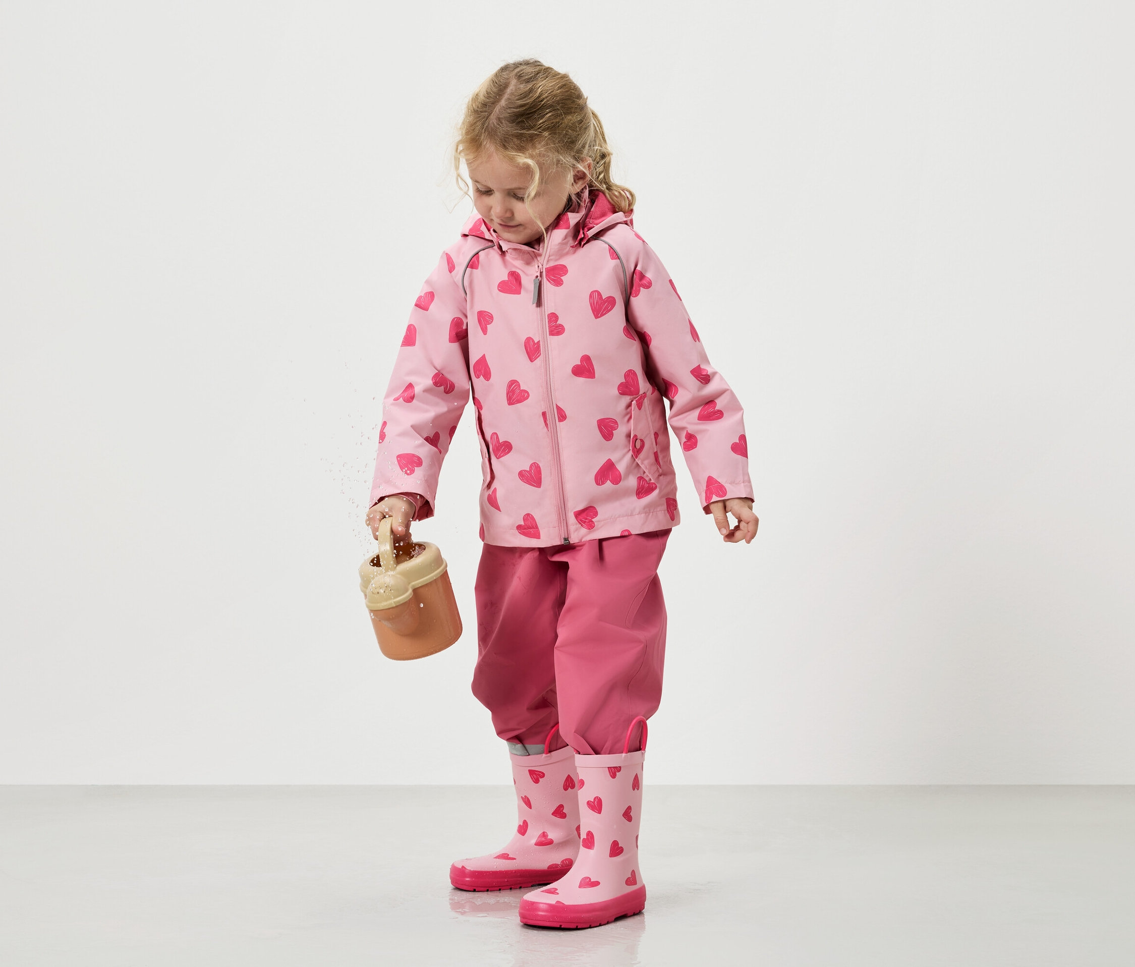 Mała dziewczynka wylewa piasek z zabawkowego wiaderka, ubrana w różową kurtkę w serduszka, pasujące spodnie i kalosze.