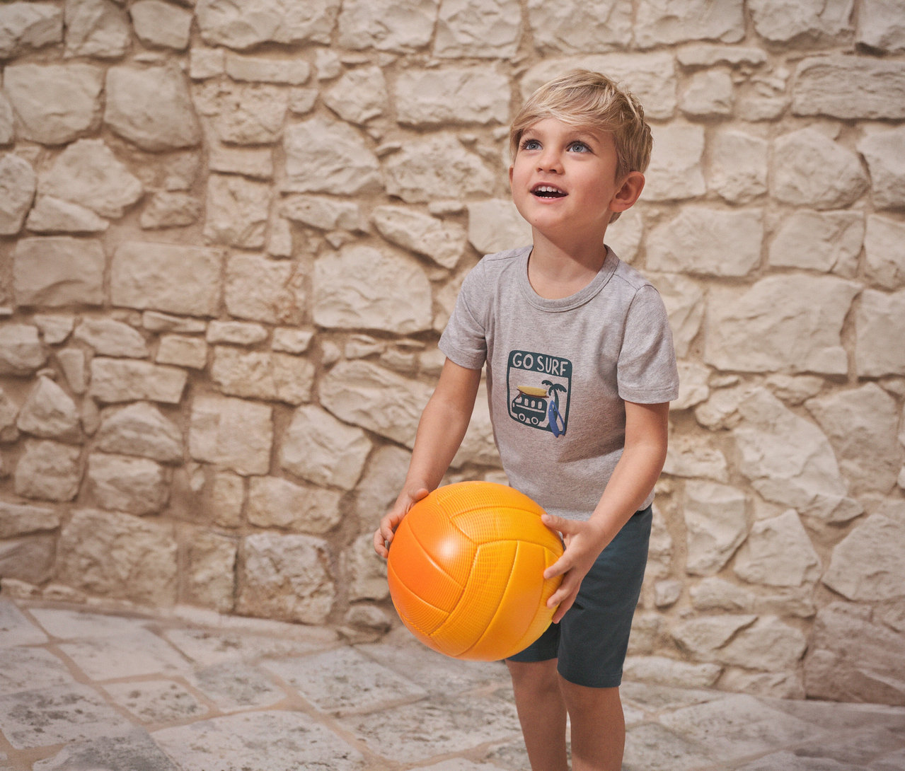 Chłopiec trzyma pomarańczową piłkę, ubrany w piżamę dziecięcą SCHIESSER na tle kamiennej ściany.