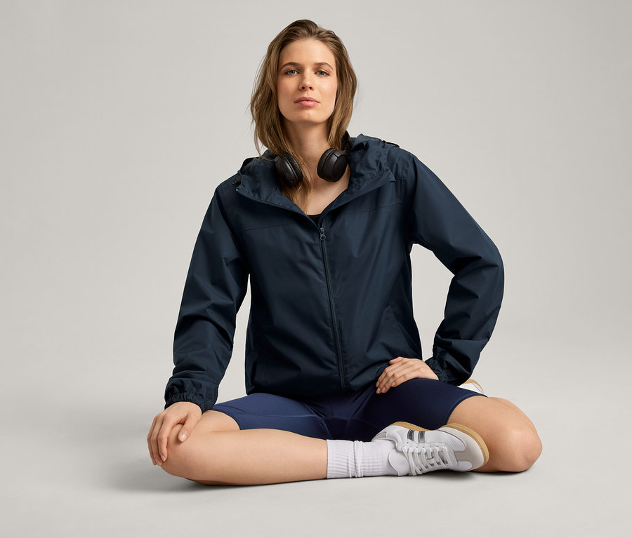 Kobieta siedzi po turecku w kurtce przeciwdeszczowej, słuchawkach, szortach i butach sportowych.