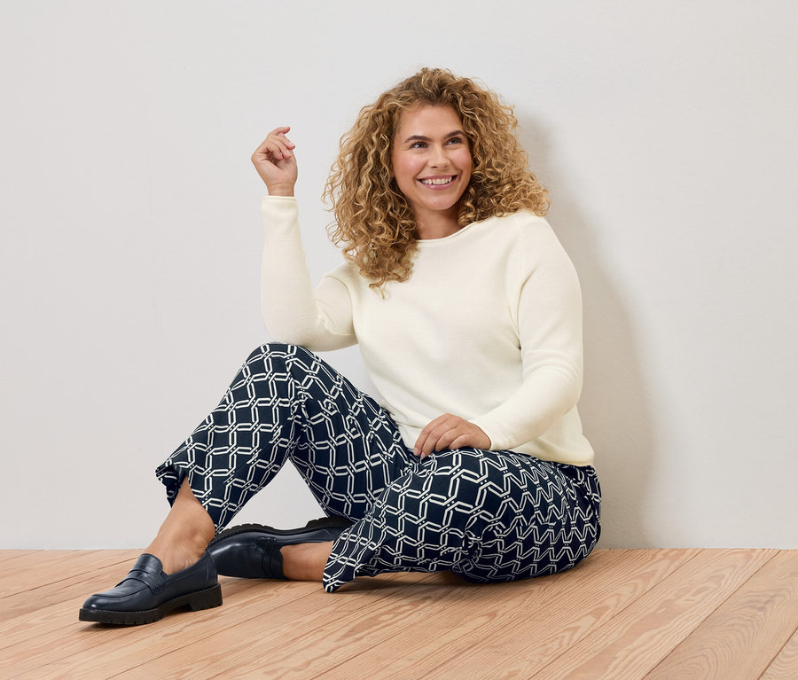 Kobieta z kręconymi włosami siedzi na drewnianej podłodze, ma na sobie kremowy sweter i niebieskie spodnie w geometryczne wzory.