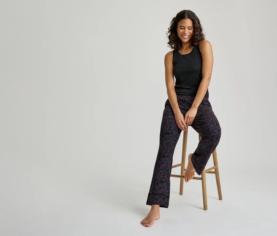 Uśmiechnięta kobieta z ciemnymi kręconymi włosami siedzi na drewnianym stołku. Ma na sobie czarny podkoszulek i ciemne wzorzyste spodnie od piżamy. Jej stopy są bose.
