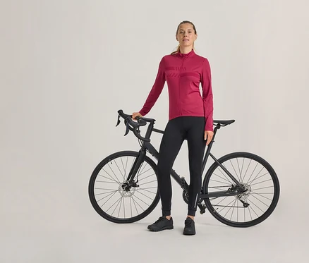 Kobieta stoi obok czarnego roweru szosowego, ubrana w fuksjową bluzę rowerową z długim rękawem, czarne spodnie rowerowe i czarne buty.