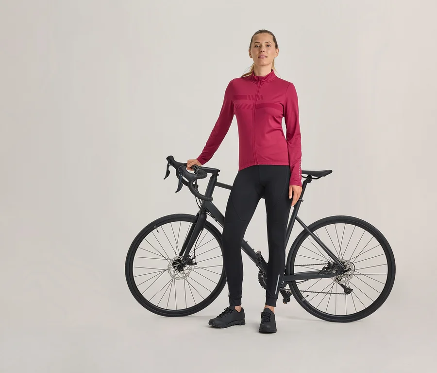 Kobieta stoi obok czarnego roweru szosowego, ubrana w fuksjową bluzę rowerową z długim rękawem, czarne spodnie rowerowe i czarne buty.