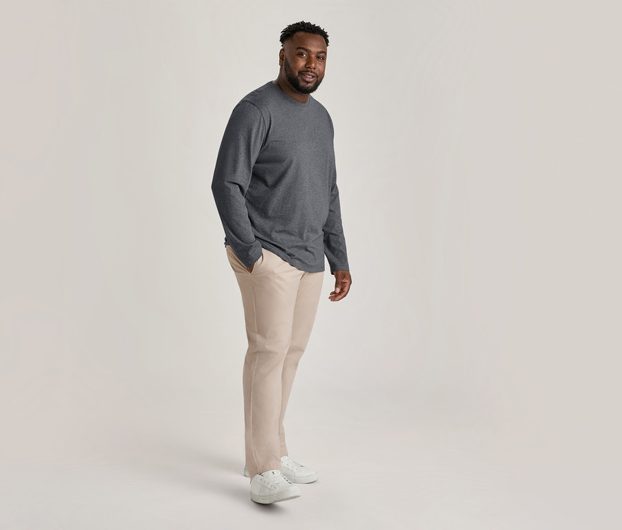 Uśmiechnięty mężczyzna w ciemnoszarym longsleeve'ie, beżowych spodniach chinosach i białych sneakersach, z jedną ręką w kieszeni.