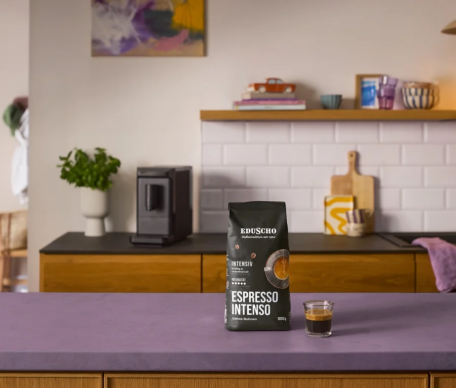 Czarna paczka kawy ziarnistej Eduscho Espresso Intenso i espresso na fioletowym blacie, za nimi czarny ekspres »Esperto mini«.
