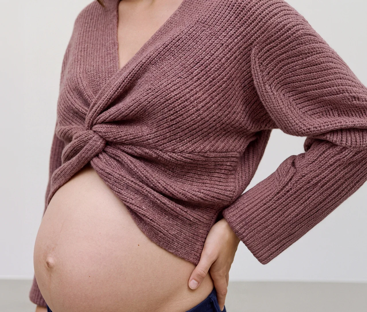 Kobieta w ciąży nosi różowy krótki top z dzianiny MAMALICIOUS „SVALA” z wiązaniem z przodu, odsłaniający brzuch. Jej lewa ręka spoczywa na biodrze.