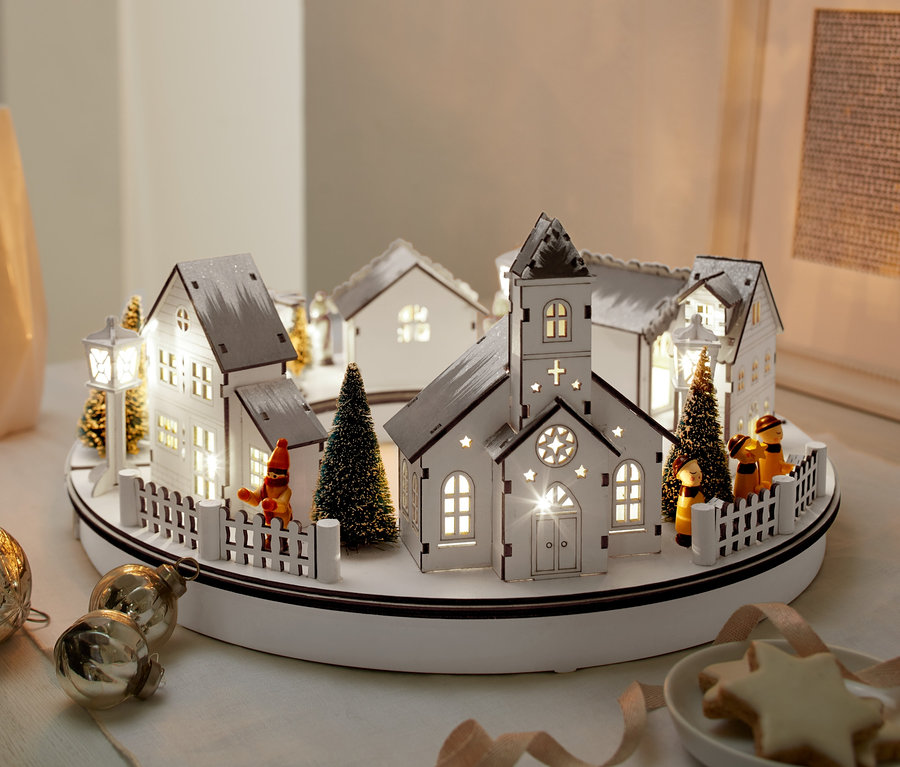 Podświetlana drewniana wioska LED obok wazonów, ozdób i talerza ciasteczek.