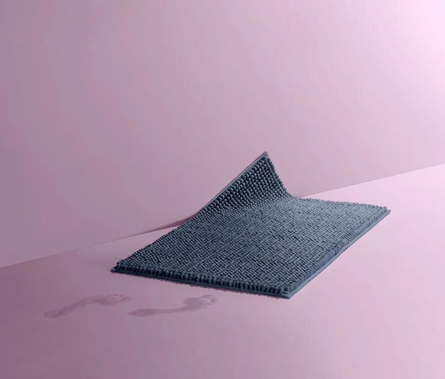 Niebieska mata łazienkowa o wymiarach ok. 50 x 80 cm na różowym tle. Na podłodze widoczne mokre ślady stóp.
