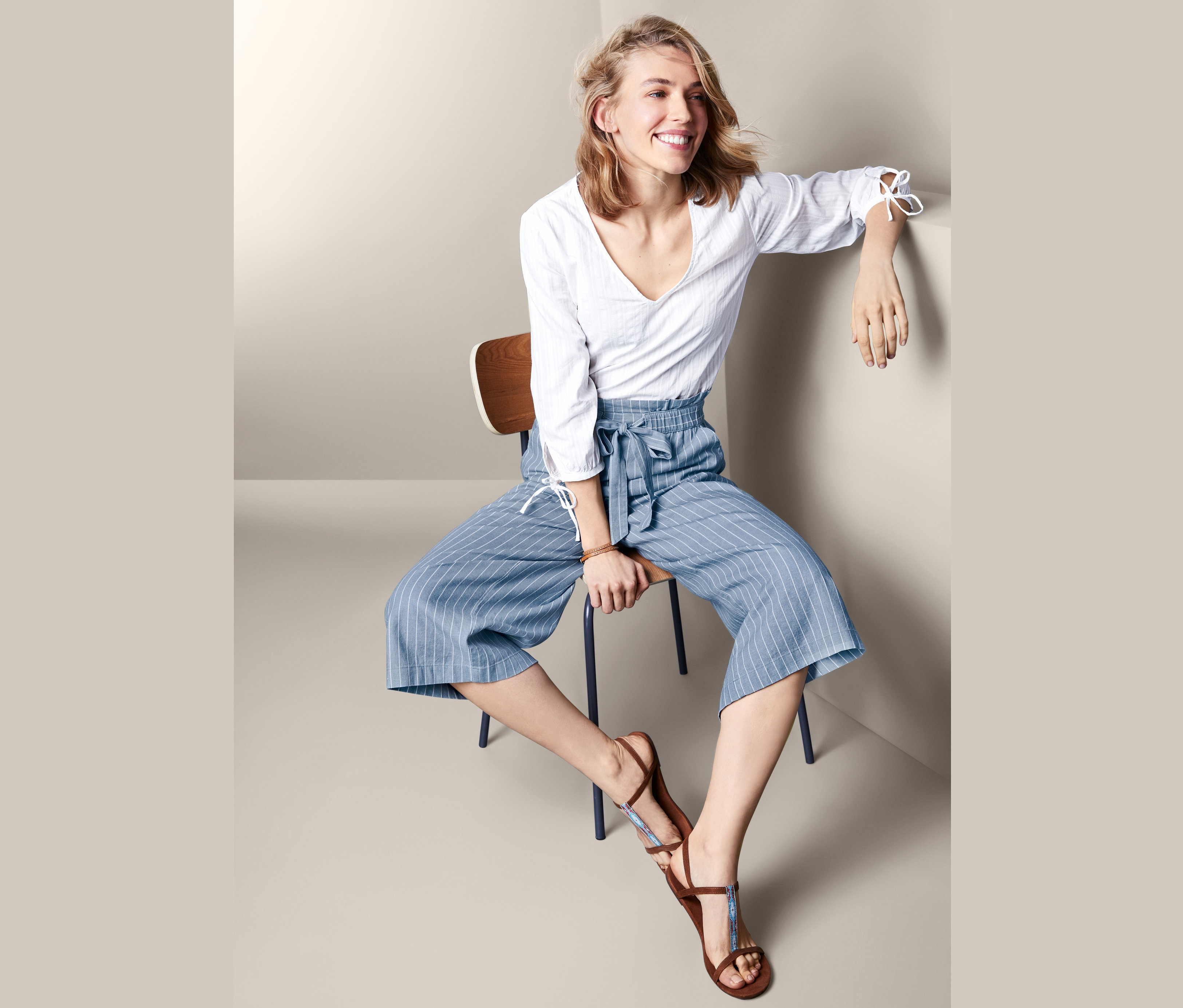 Kobieta siedzi na krześle i ma na sobie damskie spodnie kuloty z lnu i wiskozy w niebiesko-białe paski oraz białą bluzkę.
