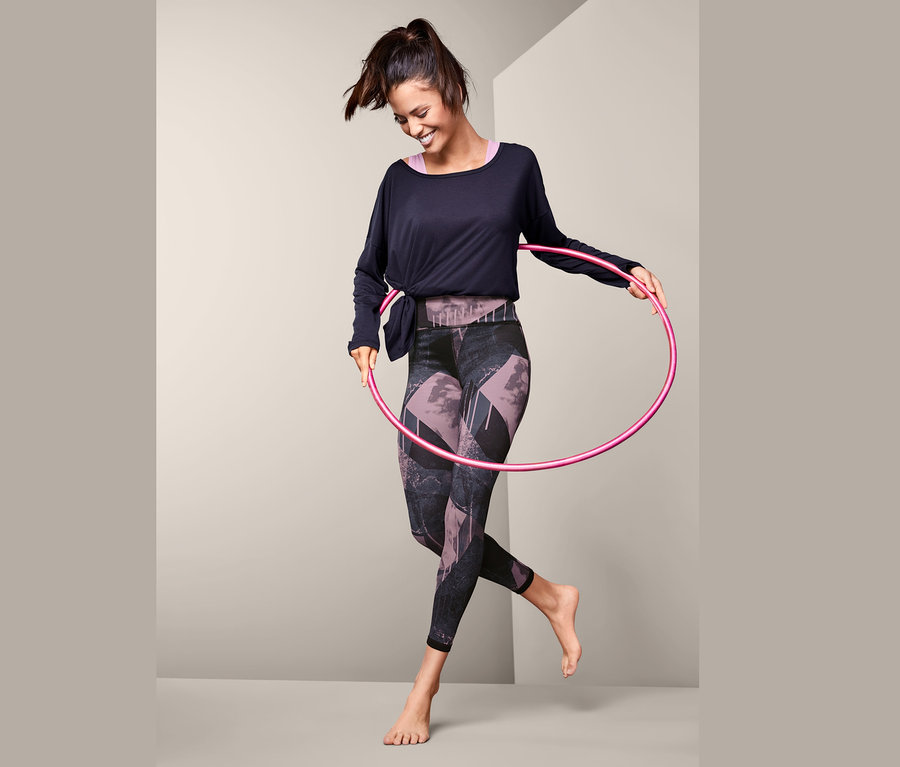 Uśmiechnięta kobieta kręci różowym hula-hoop na biodrach, ma na sobie dwustronne legginsy sportowe.