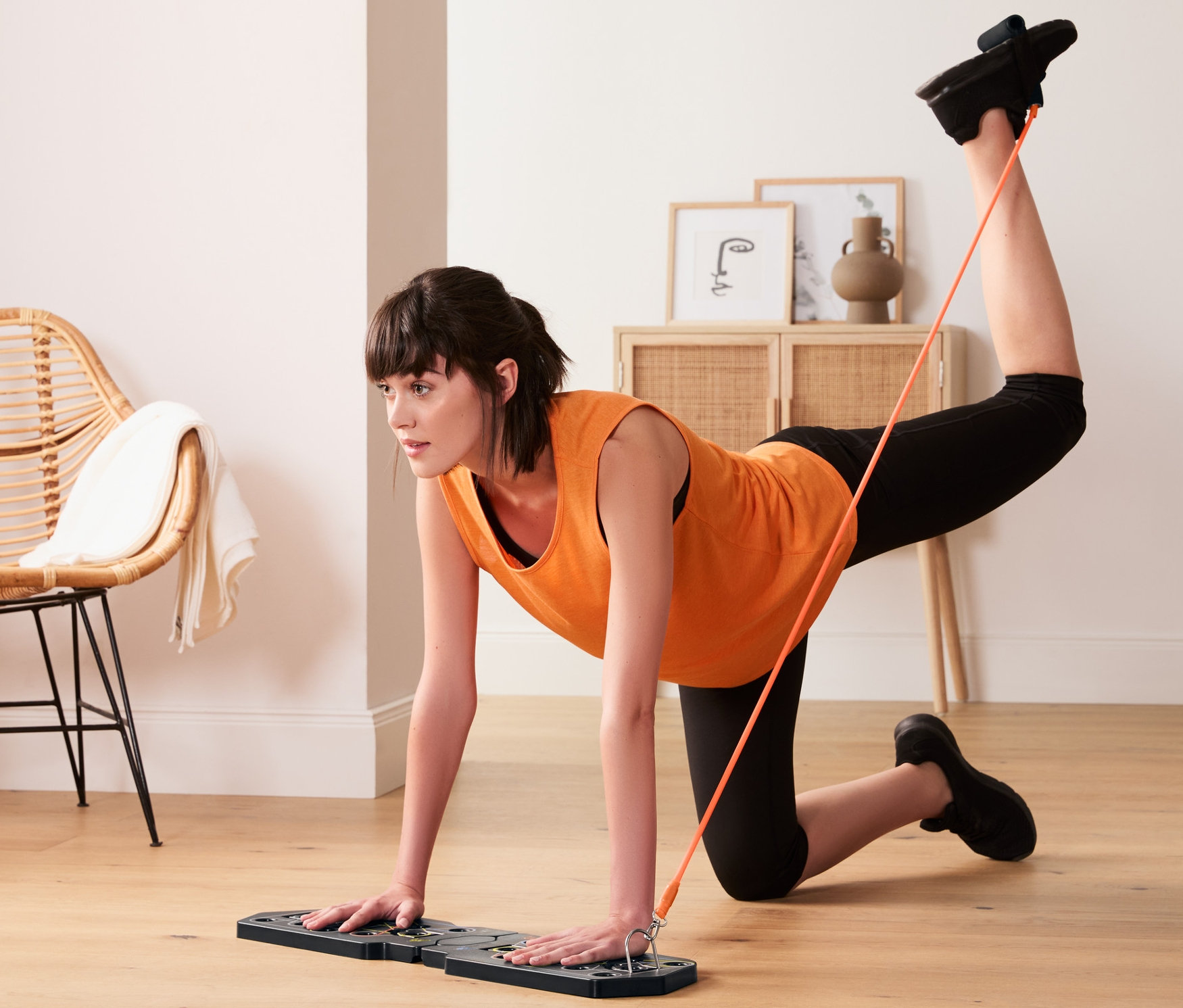 Kobieta ćwiczy z deską do pompek i z gumami do ćwiczeń, jedną ręką podpiera się na desce, a drugą nogą ciągnie gumę.