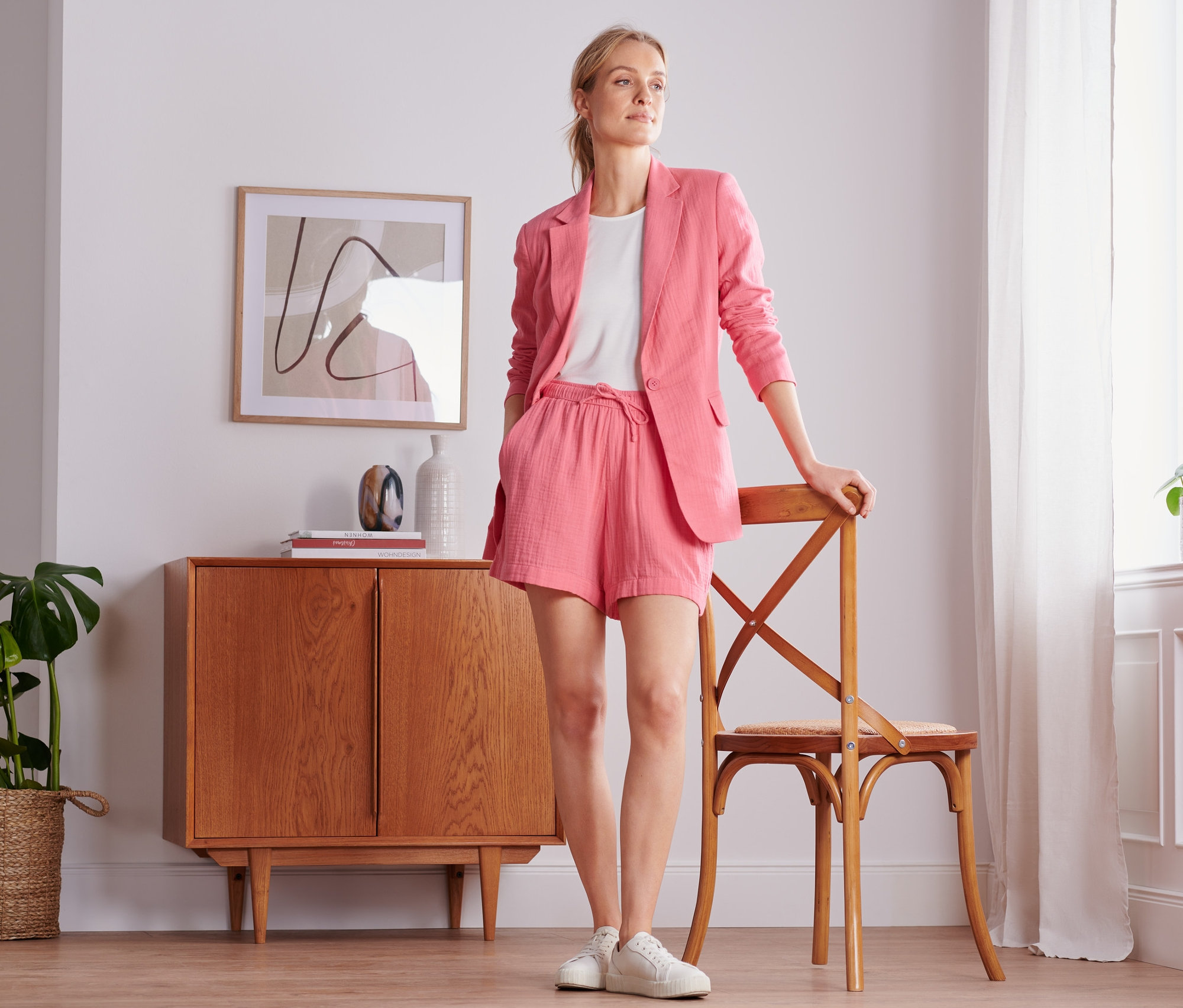Kobieta pozuje w koralowym żakiecie z muślinu i szortach. Opiera się o krzesło obok okna.