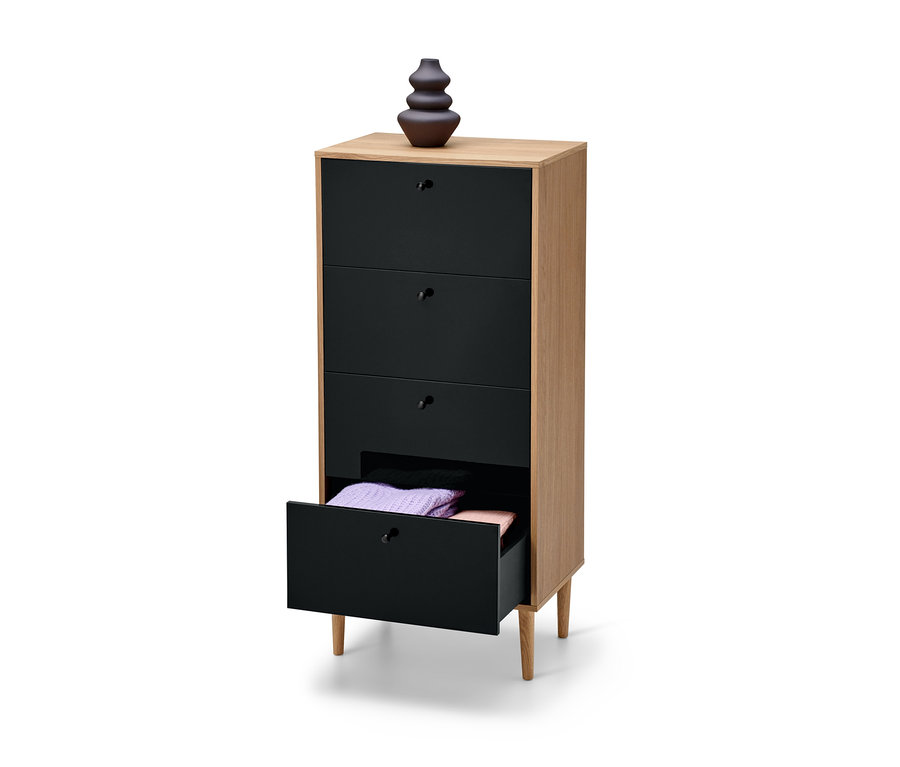 Wąska komoda »Bekeson« w kolorze antracytowym z czterema szufladami, z których jedna jest otwarta i zawiera złożone swetry. Na komodzie stoi brązowy wazon.