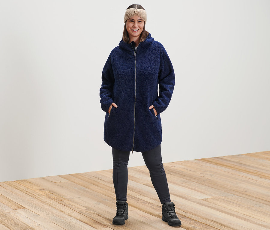 Kobieta stoi, ma na sobie płaszcz pluszowy z kapturem, opaskę z dzianiny w kolorze piaskowym i legginsy termoaktywne.