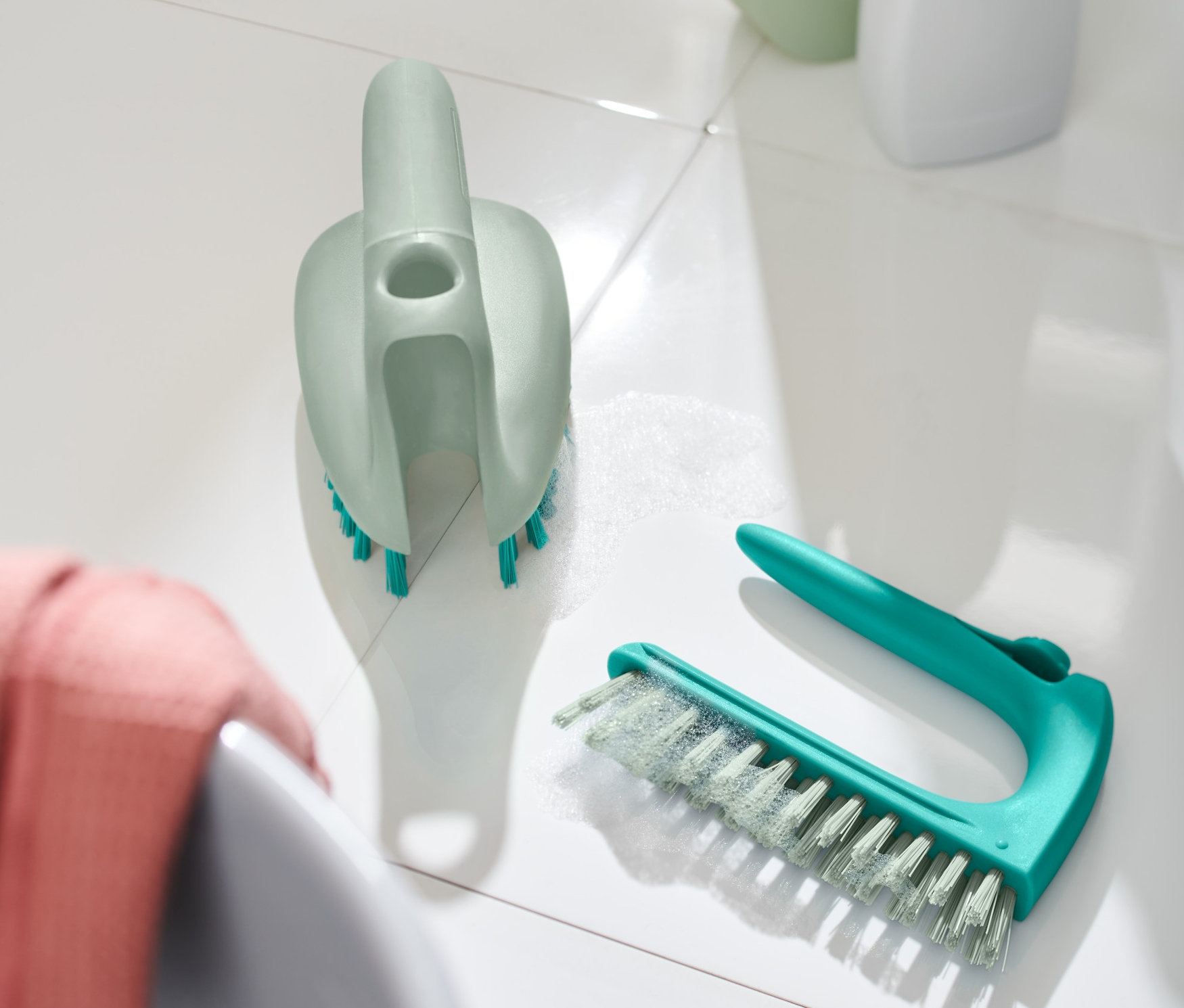 Dwie szczotki do posadzek z płytek 2 w 1 leżą na białych płytkach obok umywalki.