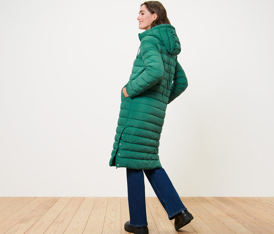 Kobieta pozuje bokiem w długim pikowanym płaszczu z kapturem.