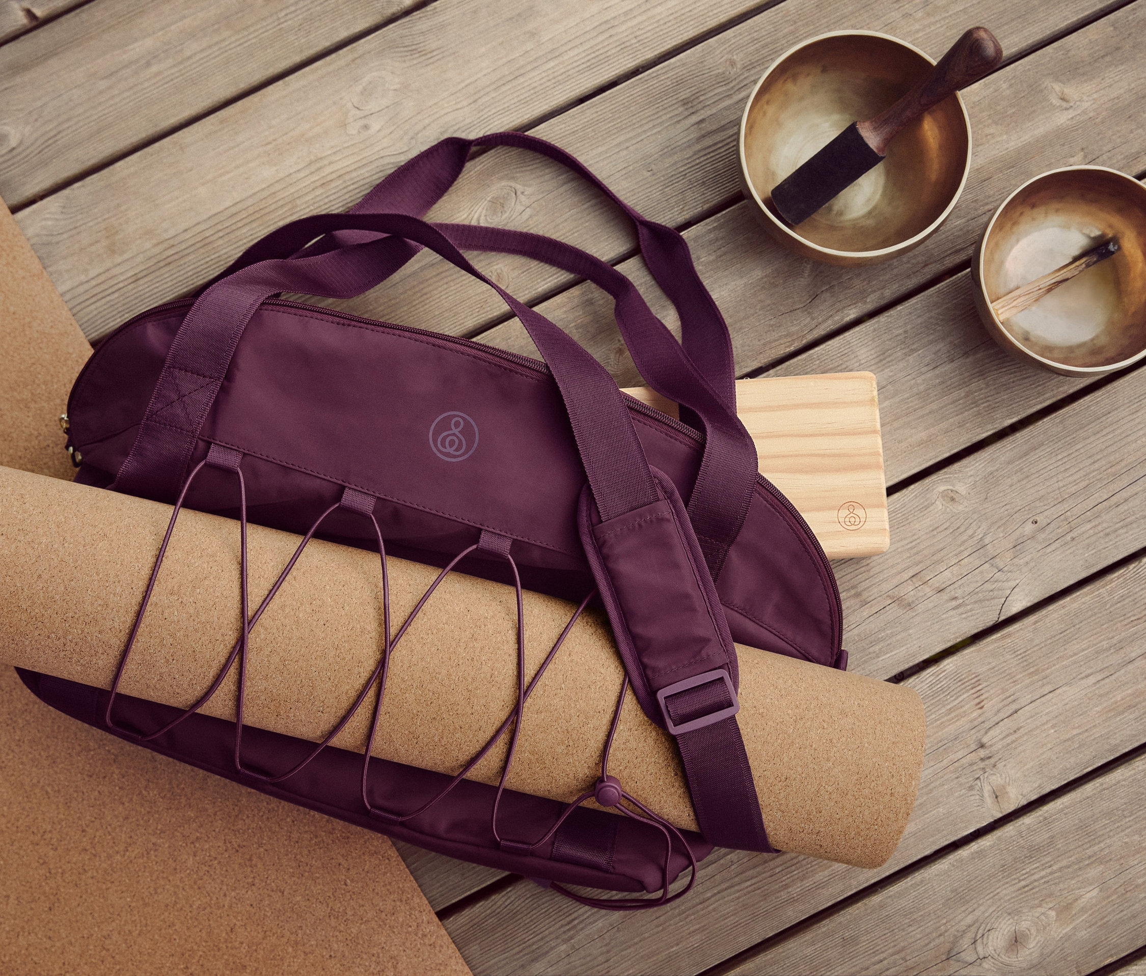 Na drewnianej podłodze leżą korkowa mata do jogi i fitnessu, torba sportowa, klocek do jogi i dwie misy tybetańskie.