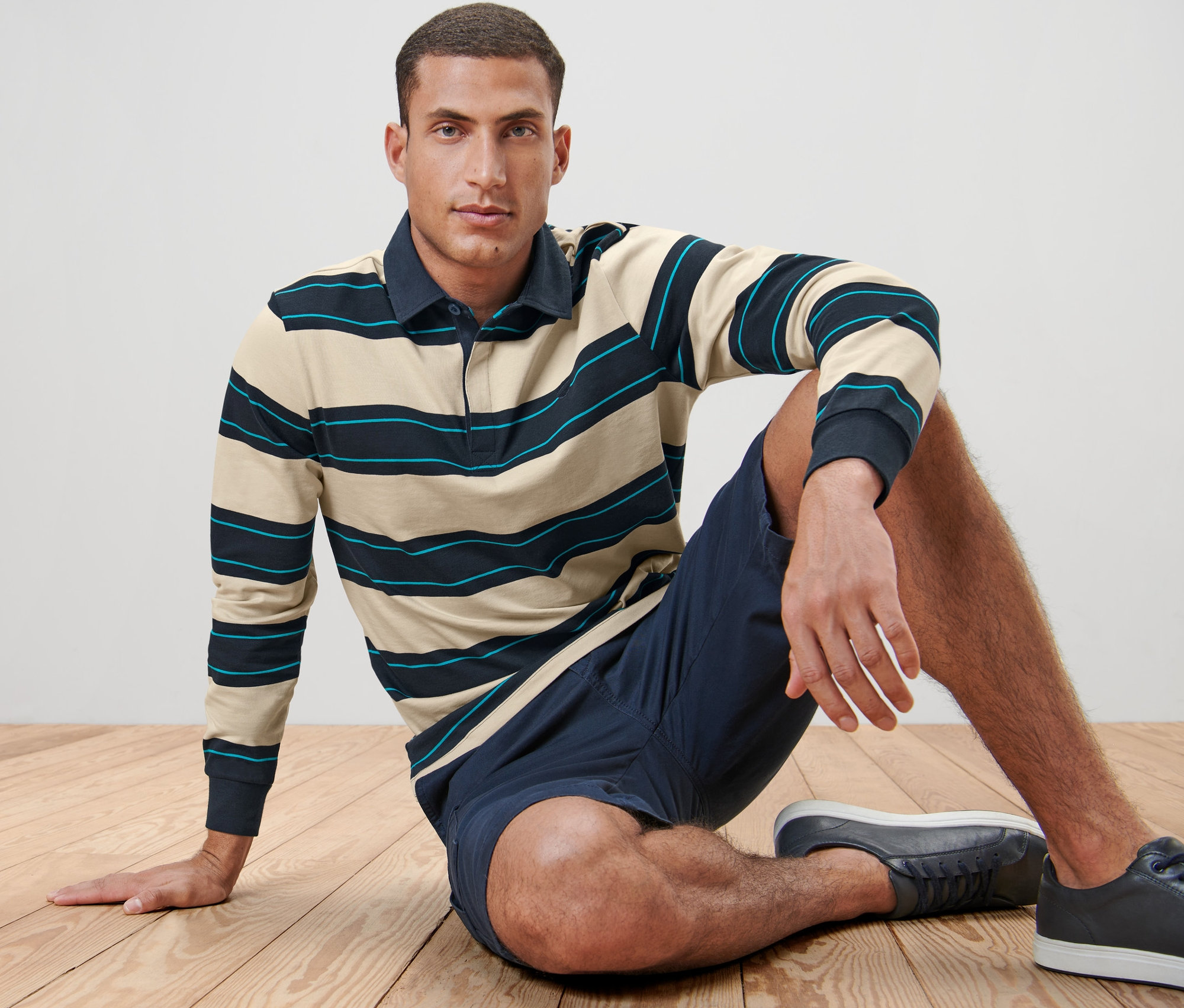 Mężczyzna siedzi na drewnianej podłodze w koszulce polo z długim rękawem, szortach i butach sportowych.