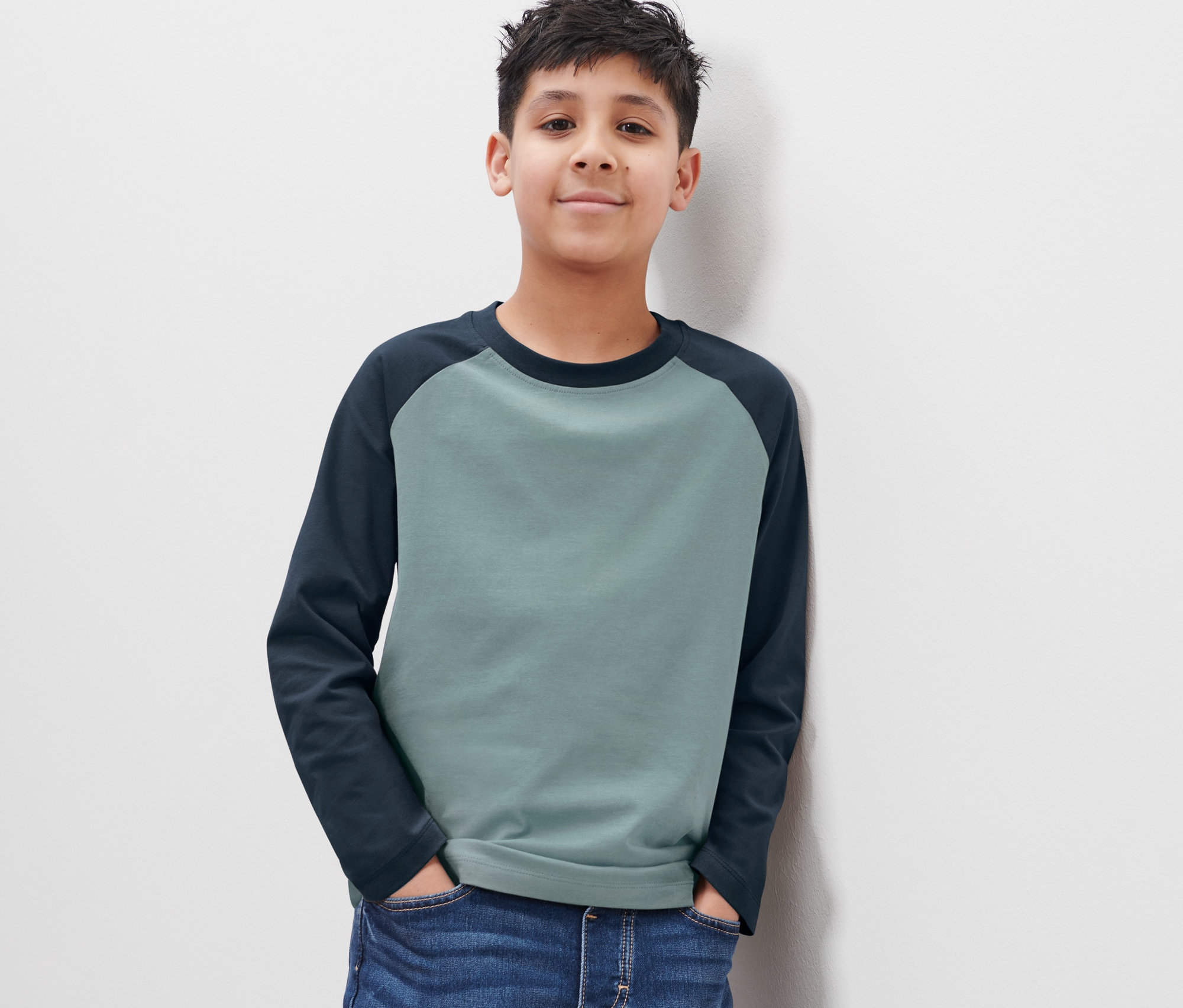 Chłopiec w niebieskich dżinsach i dwukolorowej koszulce z długim rękawem stoi tyłem do białej ściany.