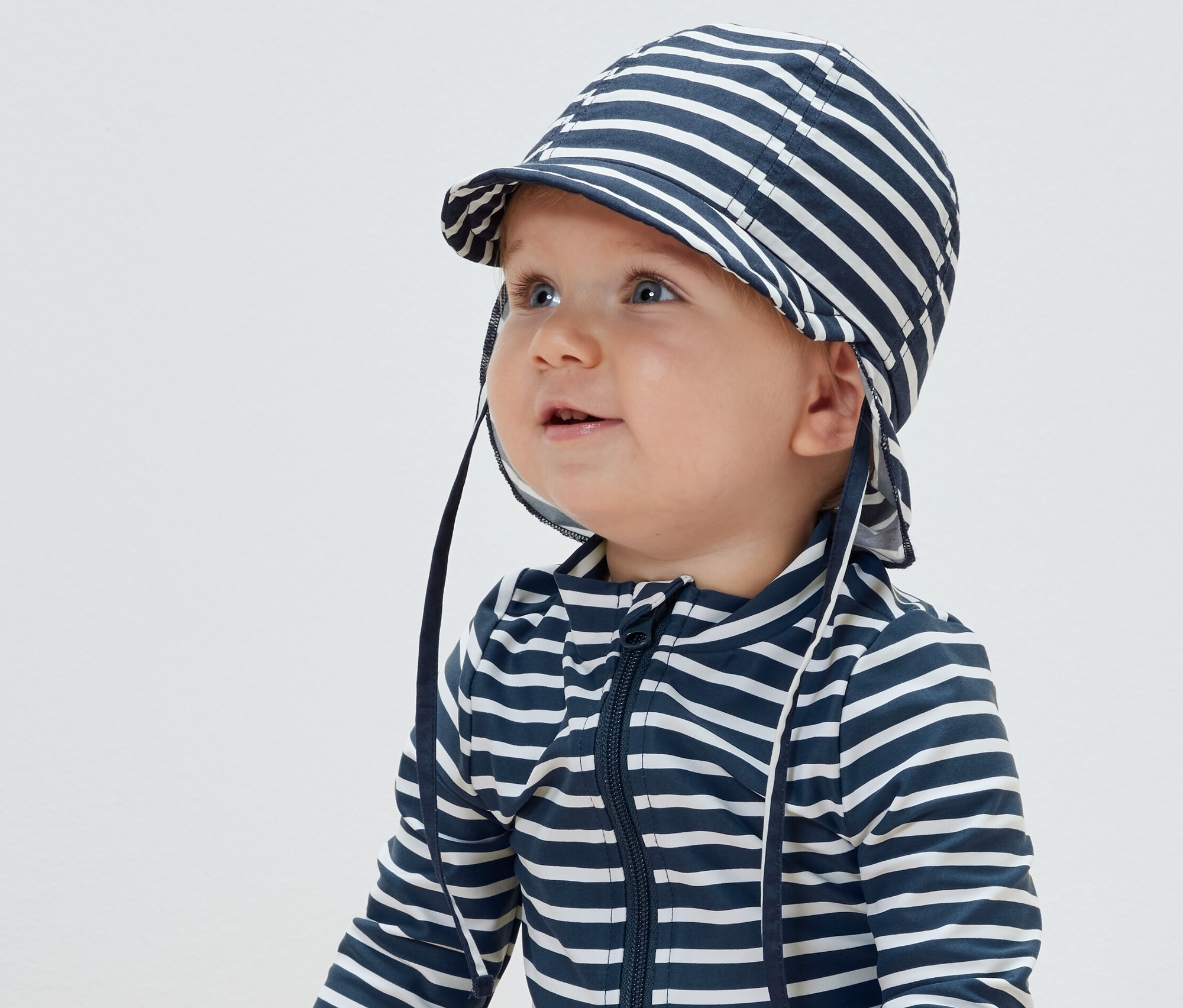 Dziecko ma na sobie czapkę przeciwsłoneczną w paski z filtrem UV 80 i kombinezon w paski.