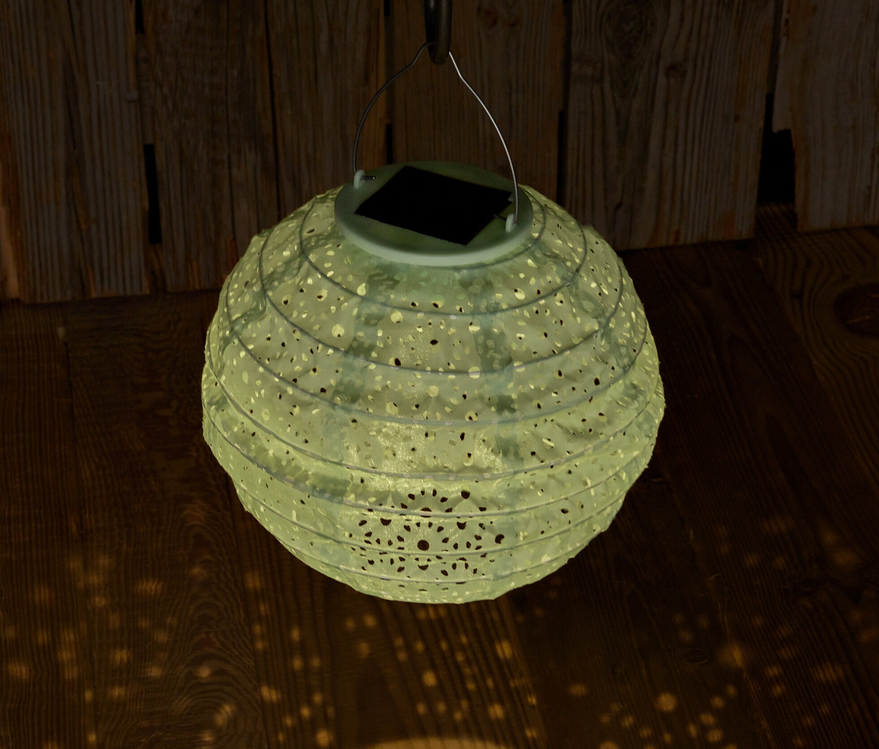 Wiszący lampion solarny z ozdobnymi otworami rzuca światło na drewnianą podłogę.