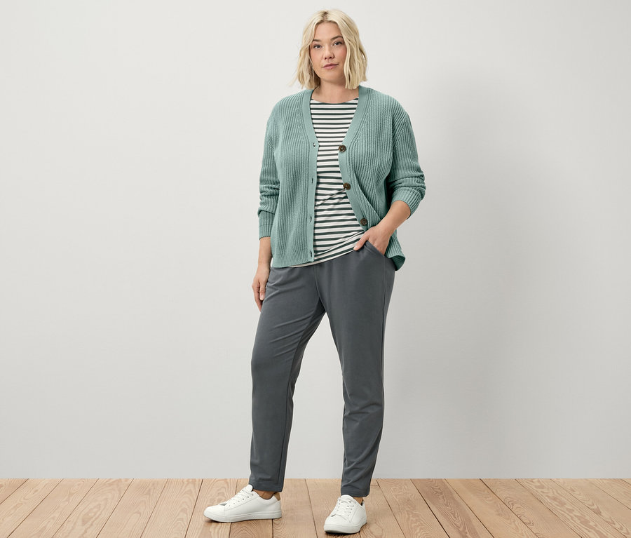 Kobieta stoi ubrana w spodnie joggery, szałwiowy kardigan o grubym splocie i koszulkę z półdługim rękawem w zielone paski.