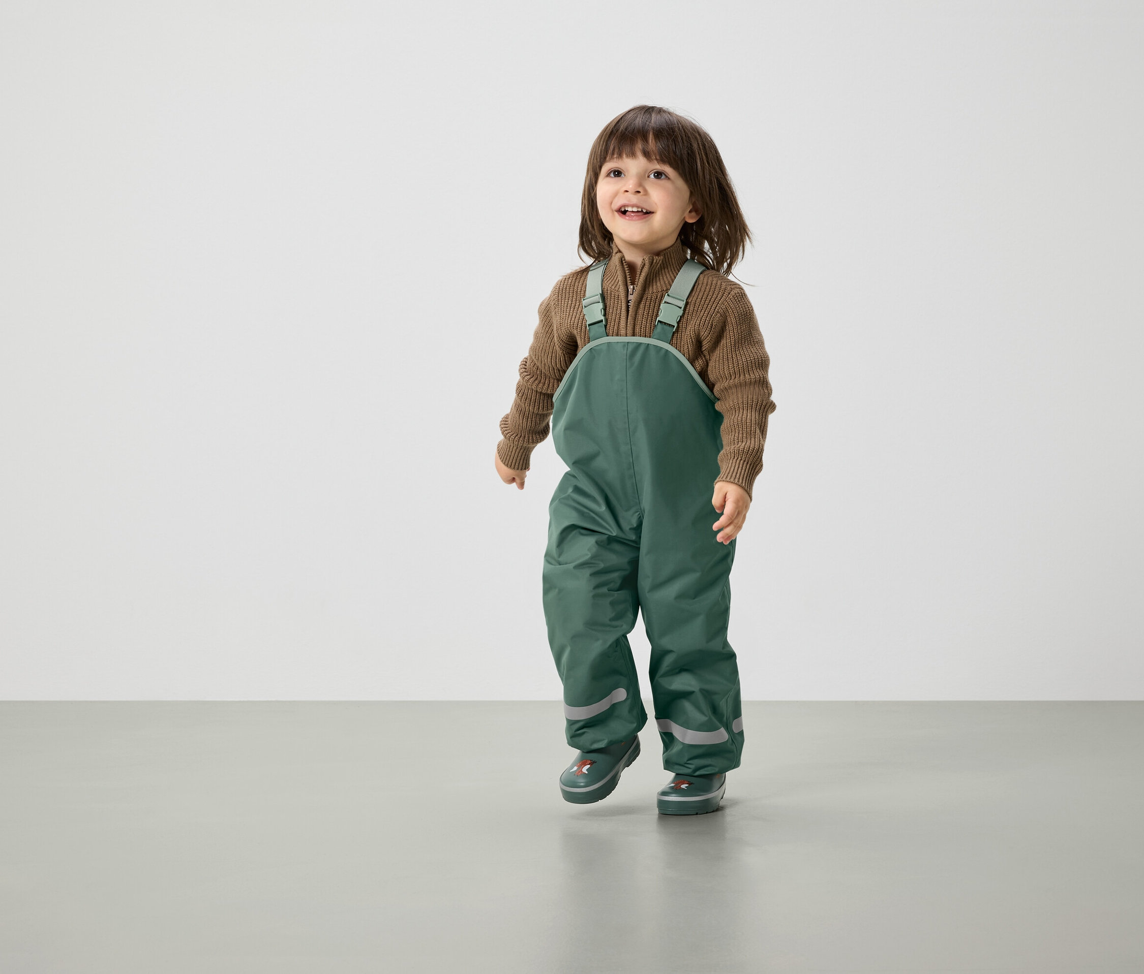 Uśmiechnięte dziecko z brązowymi włosami idzie w zielonych spodniach na szelkach i brązowym swetrze.