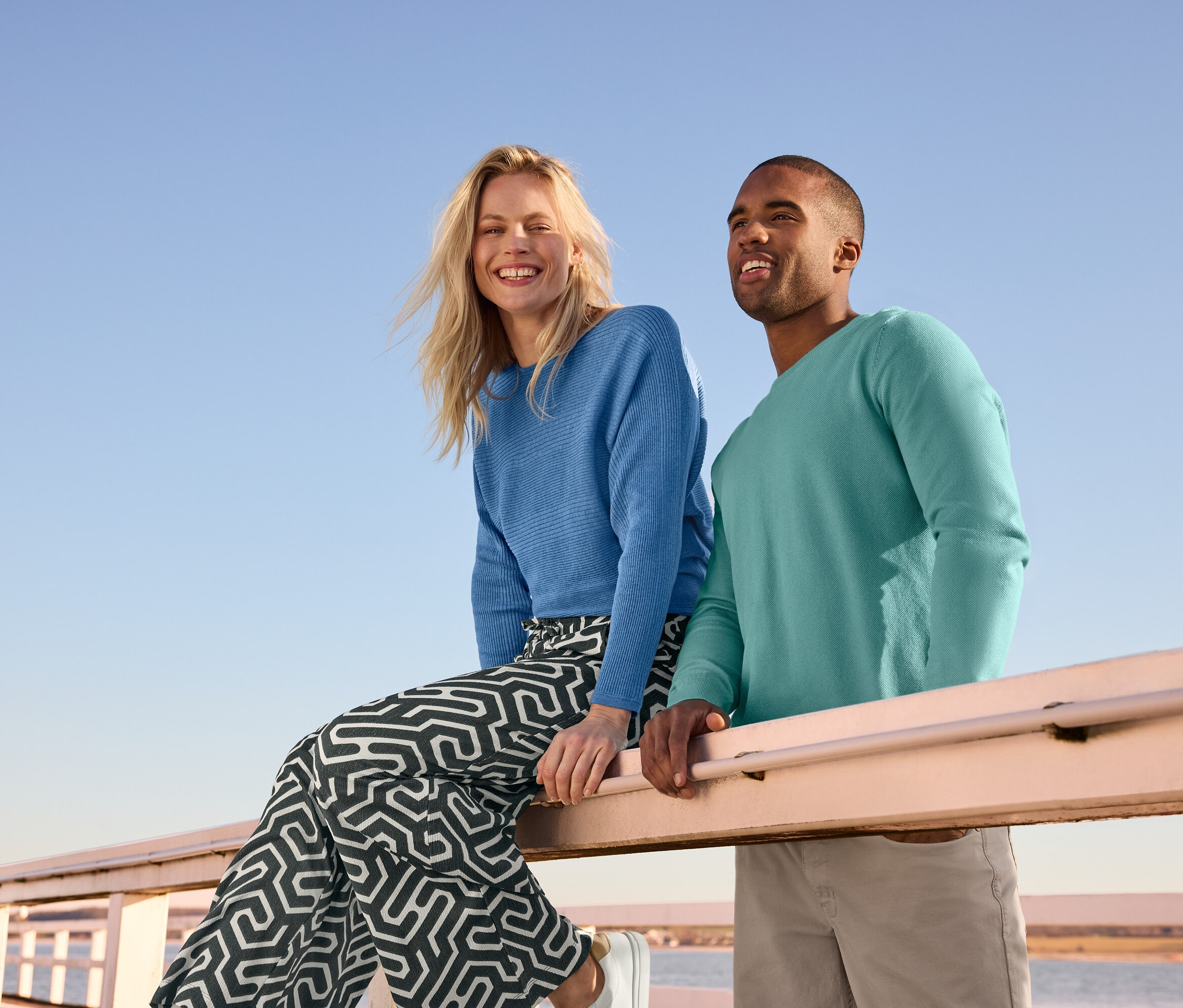 Kobieta siedzi na barierce w niebieskim, cienkim swetrze i spodniach na gumce z nadrukiem i efektem marszczenia. Obok stoi mężczyzna w swetrze i spodniach.