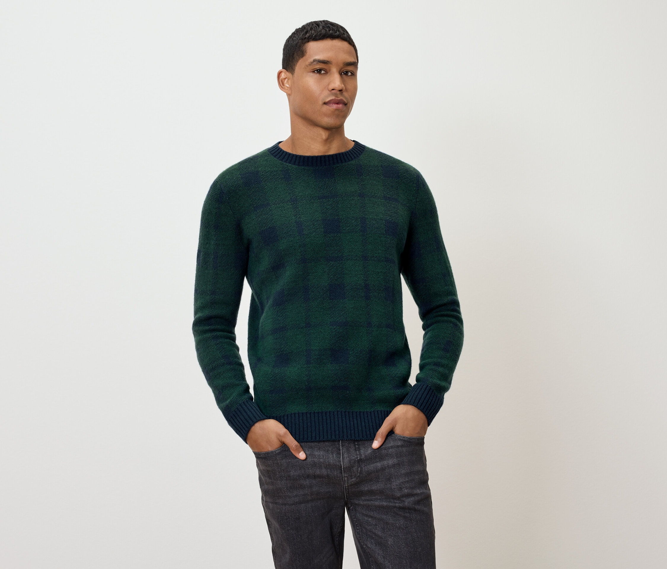 Mężczyzna pozuje w zielonym swetrze w kratę i szarych dżinsach.