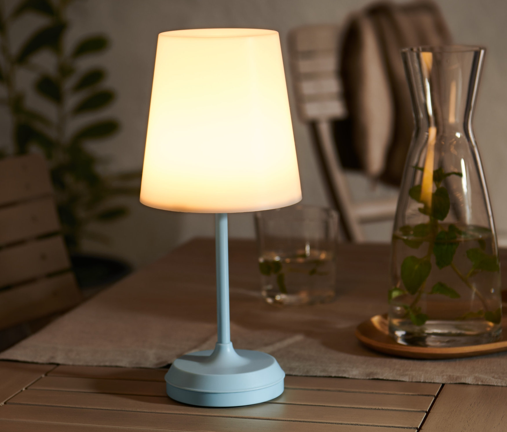 Na drewnianym stole stoi akumulatorowa lampa LED, dzbanek z wodą i szklanki.