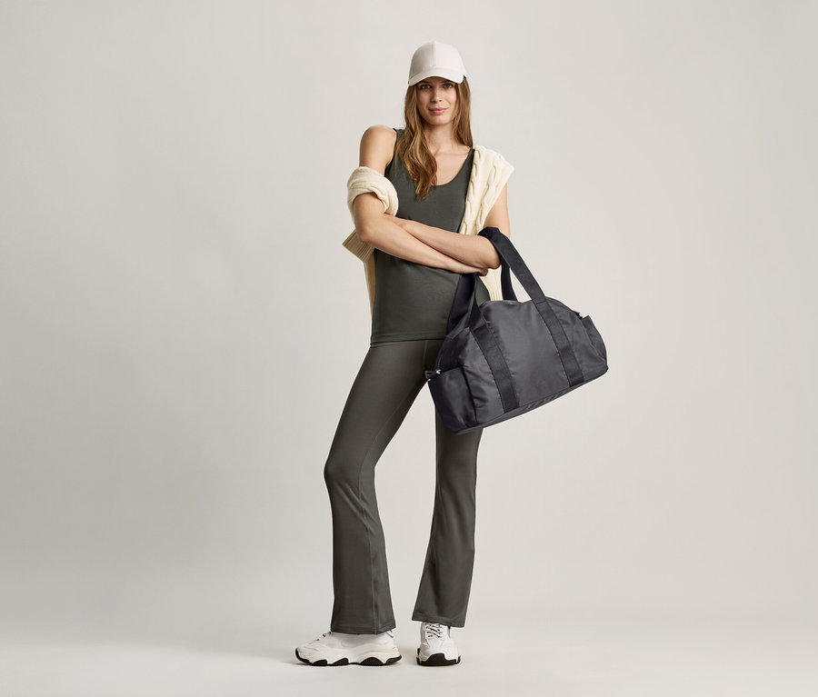 Kobieta stoi ze skrzyżowanymi ramionami, trzyma czarną torbę sportową. Ma na sobie oliwkowy top i szare spodnie.